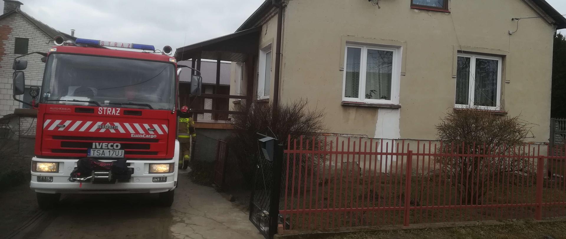 Zdjęcie przedstawia budynek jednorodzinny. Na podwórku przed domem e widać samochód ratowniczo- gaśniczy i strażaka idącego w kierunku kotłowni.