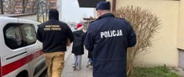 Na zdjęciu widzimy dwóch funkcjonariuszy, po lewej stronie z Państwowej Straży Pożarnej oraz po prawej stronie funkcjonariusza policji. Obaj zwróceni są tyłem. Funkcjonariusze roznoszą paczki dla kombatantów powiatu radomskiego. Po lewej stronie na zdjęciu ustawiony jest pojazd straży pożarnej w kolorze białym i czerwonym paskiem wzdłuż boku pojazdu. Strażak ubrany jest w munudr - czarny polar i beżowe spodnie, na polarze widnieje napis Państwowa Straż Pożarna koloru zółtego. Policjant również jest ubrany w mundur koloru granatowego, na którym widnieje napis Policja.