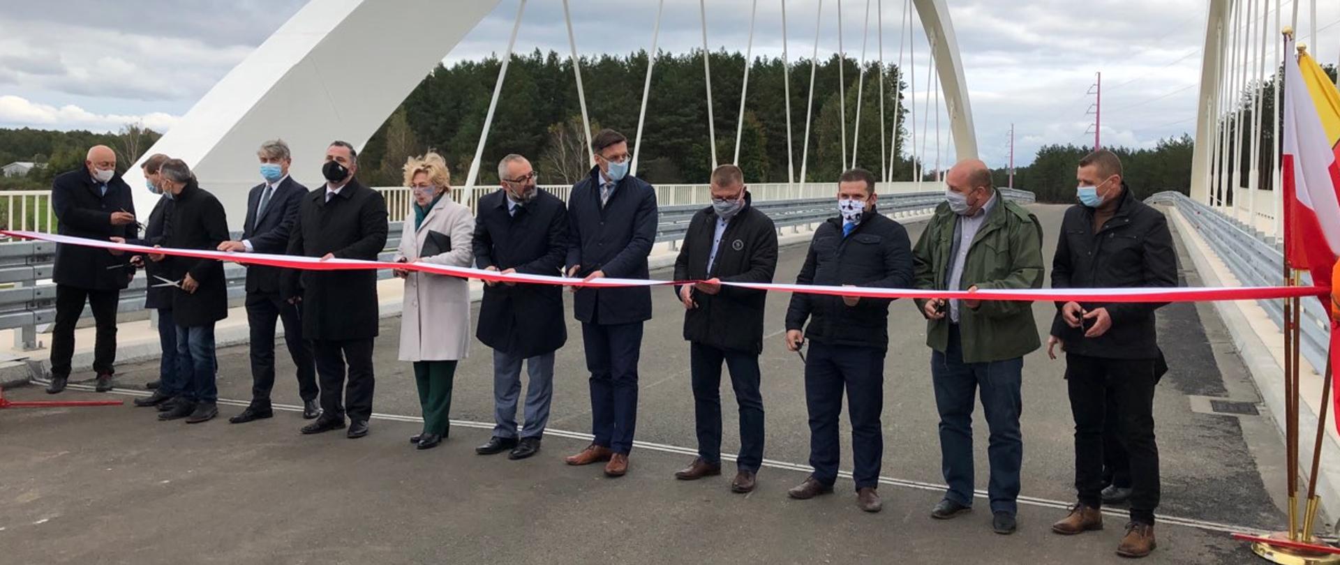 Grupa kilkunastu osób przecina nożyczkami wstęgę w kolorze biało-czerwonym dokonując symbolicznego otwarcia mostu.