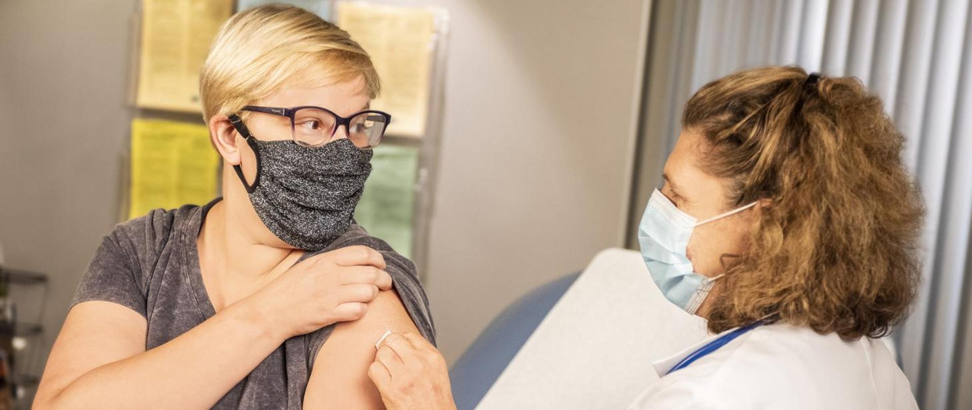 Dwie kobiety w maseczkach - pacjentka i pielęgniarka. Pacjentka podnosi rękaw w celu wykonania zabiegu szczepienia.