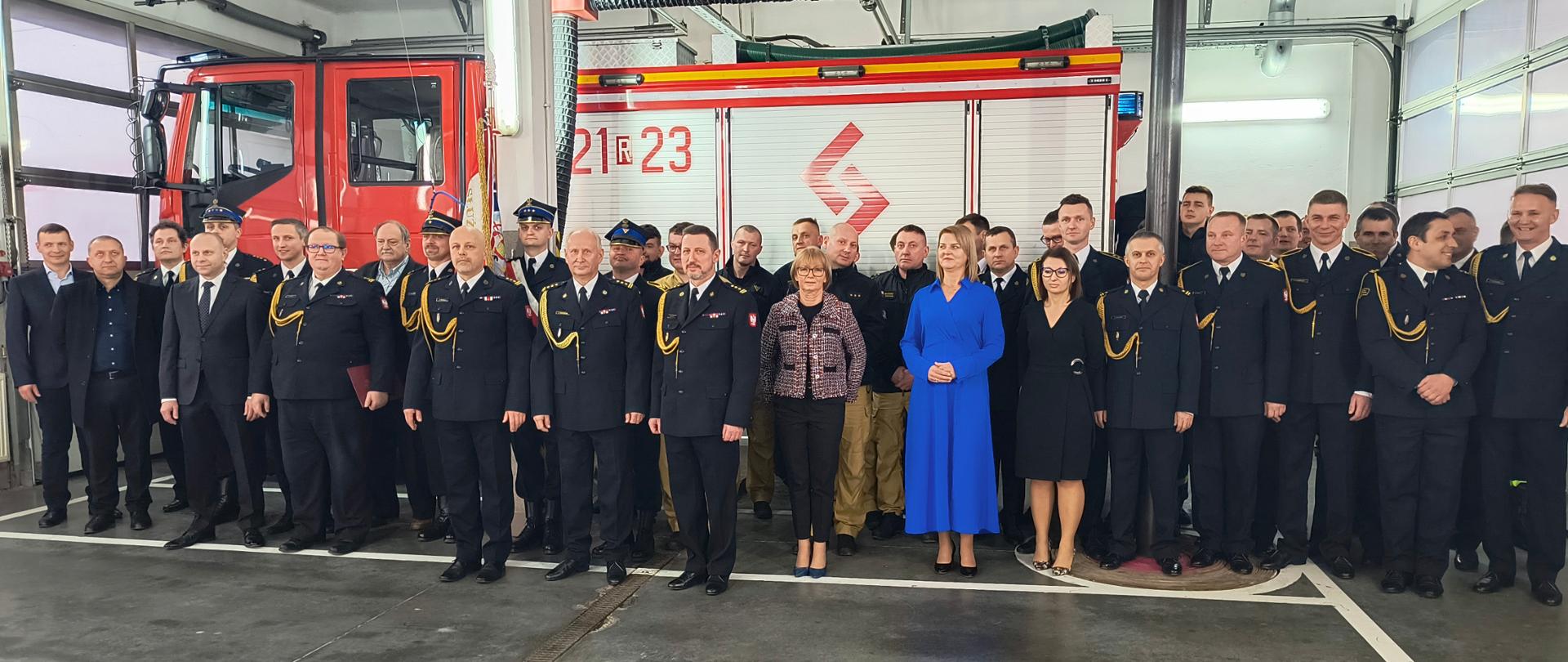 Strażacy ubrani w mundury wyjściowe i pracownicy cywilni Komendy Powiatowej Państwowej Straży Pożarnej w Dębicy stoją w garażu ustawieni w 3 rzędach przed samochodem pożarniczym. Na środku stoi dowódca jednostki, który przechodzi na emeryturę. 