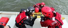 Strażacy w łódce ubrani w suche skafandry wyciągają na łódkę deskę ratowniczą z pozorantem - strażakiem. W wodzkie asekuruje strażak ubrany w suchy skafander i kask .