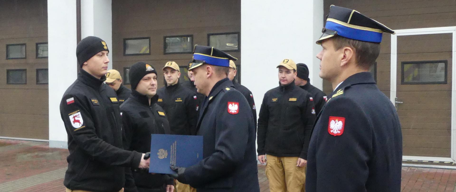 Zdjęcie przedstawia st. kpt. Krzysztofa Natuckiego - Komendanta Powiatowego PSP w Brodnicy wręczającego decyzje o awansie na wyższy stopień służbowy starszemu sekcyjnemu Sebastianowi Janc. Panowie ściskają sobie dłonie. Za komendantem stoi jego zastępca. Za dwójką awansowanych stoją strażacy podziału bojowego JRG.