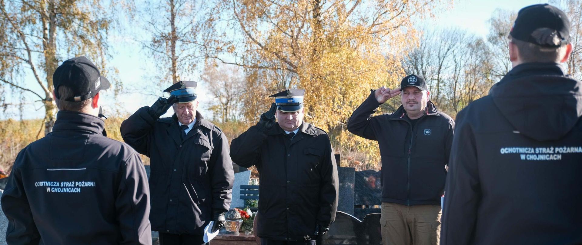 Cmentarz. Słoneczny dzień. Komendant powiatowy wraz ze strażakami salutuje przed grobem. 