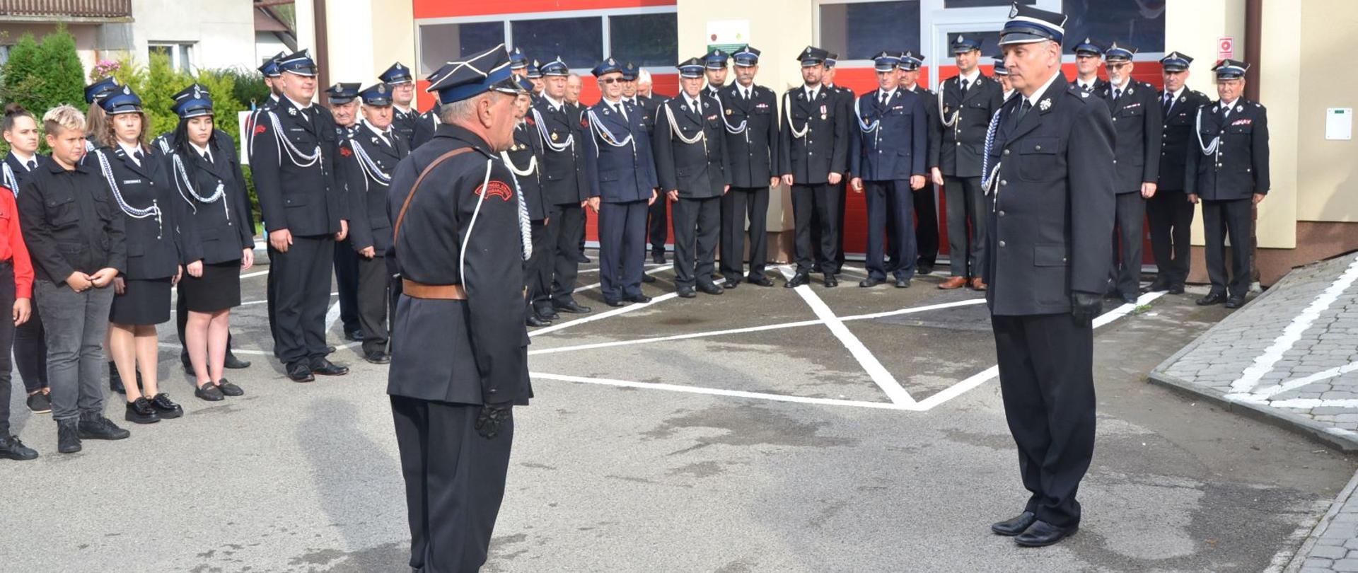 Kolorowa fotografia wykonana w pogodny dzień. Przedstawia strażaków w szyku zwartym, młodzieżową drużyną pożarniczą i dowódcą uroczystości podczas składania meldunku z okazji120 lecia OSP w Domaradzu. Meldunek odbiera Prezes Zarządu Oddziału Powiatowego ZOSP w Brzozowie. W tle bramy garażowe.