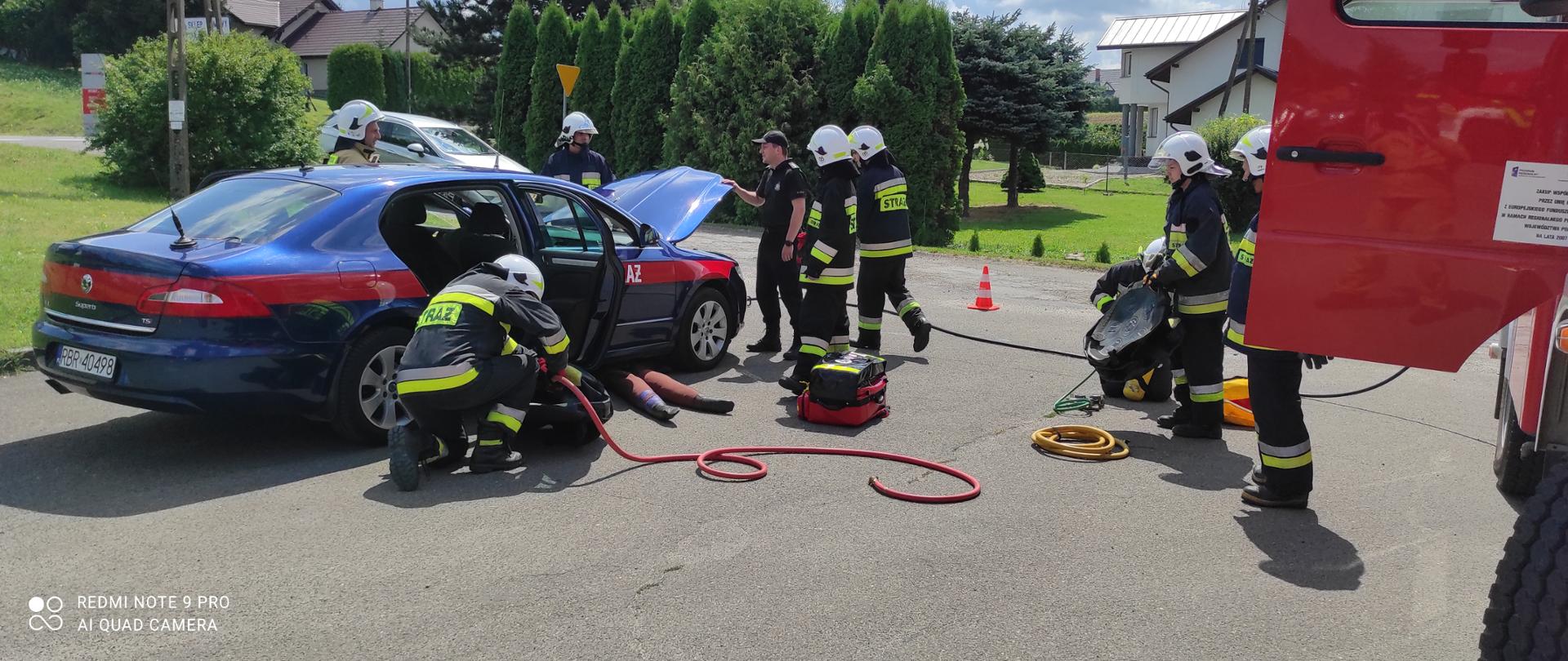 Kolorowa fotografia wykonana na zewnątrz w pogodny dzień. Przedstawia samochód strażacki osobowy z otwartymi drzwiami i maską silnika. Obok samochodu osobowego działania ratownicze prowadzą strażacy OSP ubrani w ubraniach specjalnym z hełmami na głowie. Pod samochodem znajduje się przygnieciony manekin którego mają za zadanie uwodnić strażacy.