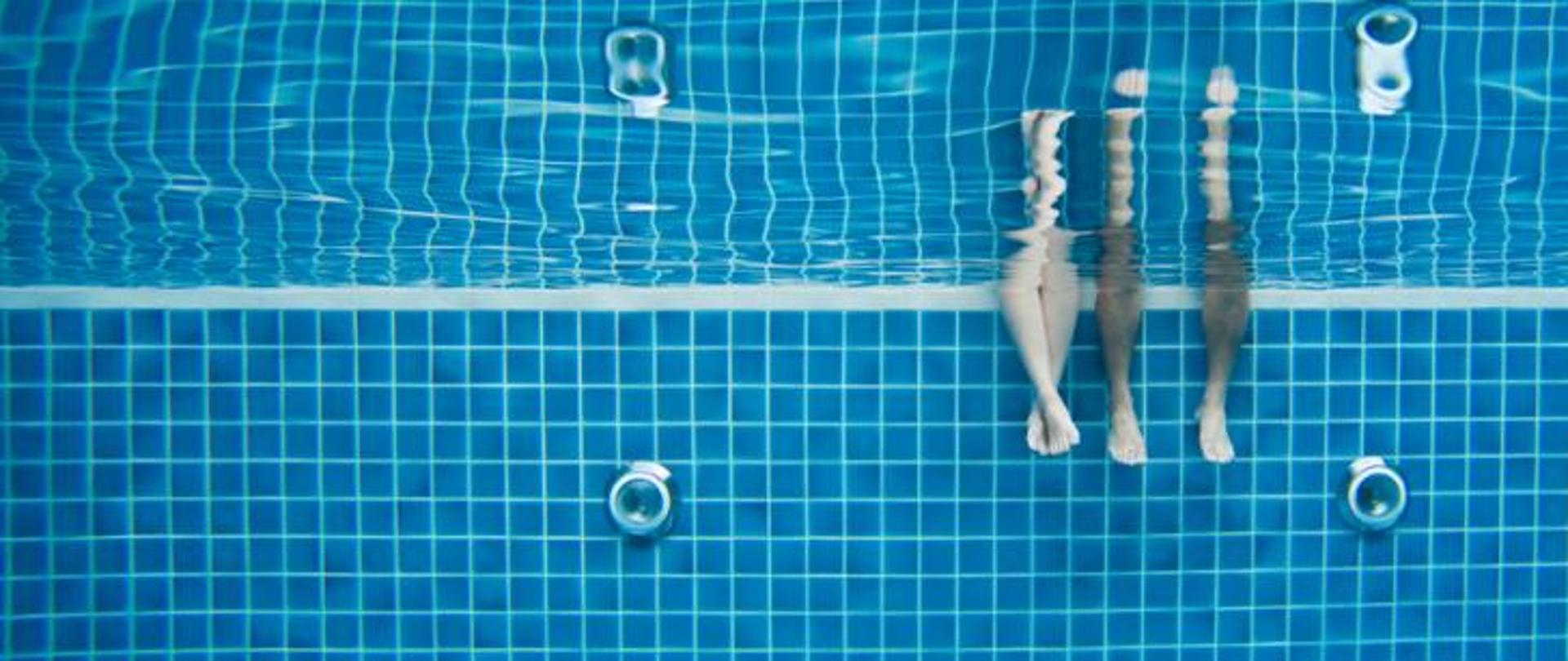 Widok basenu pod taflą wody. Po prawej stronie widać dwie pary nóg zanurzonych w wodzie.