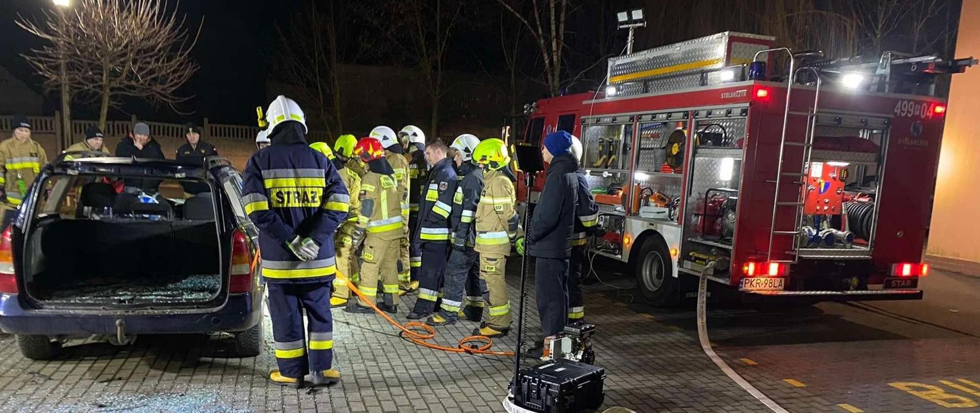 Na pierwszym planie sprzęt ratownictwa technicznego wykorzystywany do ćwiczeń rozłożony na płachcie. Druhowie ćwiczą techniki cięcia na pojeździe. Obok pojazdu samochód pożarniczy ze sprawionym oświetleniem zewnętrznym. 