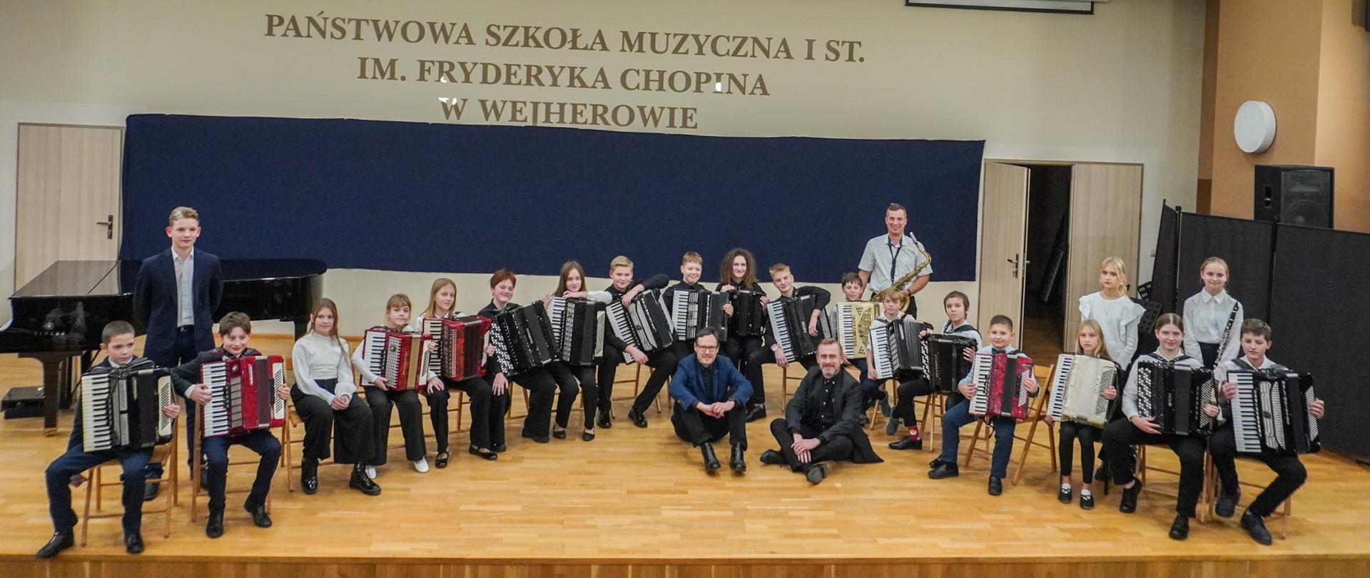 Na sali koncertowej wejherowskiej szkoły muzycznej pozują do zdjęcia wszyscy uczniowie klasy akordeonu Andrzeja Formeli i Ryszarda Borysionka. Uczniowie siedzą z akordeonami w półokręgu na krzesłach, przed nimi siedzą na scenie obaj nauczyciele. 