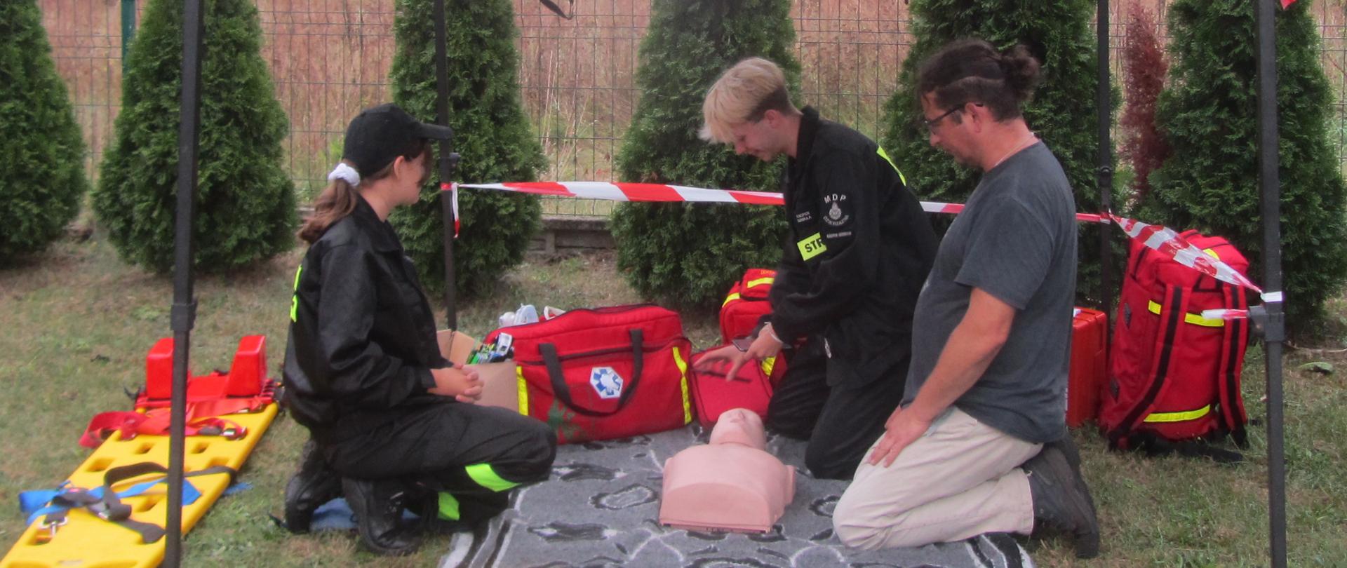 Dwoje młodych ludzi w strojach strażaków klęczy na macie, pomiędzy nimi leży fantom człowieka, obok stoją torby i plecaki z zestawami ratownictwa. Obok klęczy mężczyzna – uczestnik pokazu.