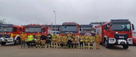 Na pierwszym planie widać strażaków i pracowników firmy ustawionych w szeregu, zdjęcie grupowe. W tle stoją samochody pożarnicze.