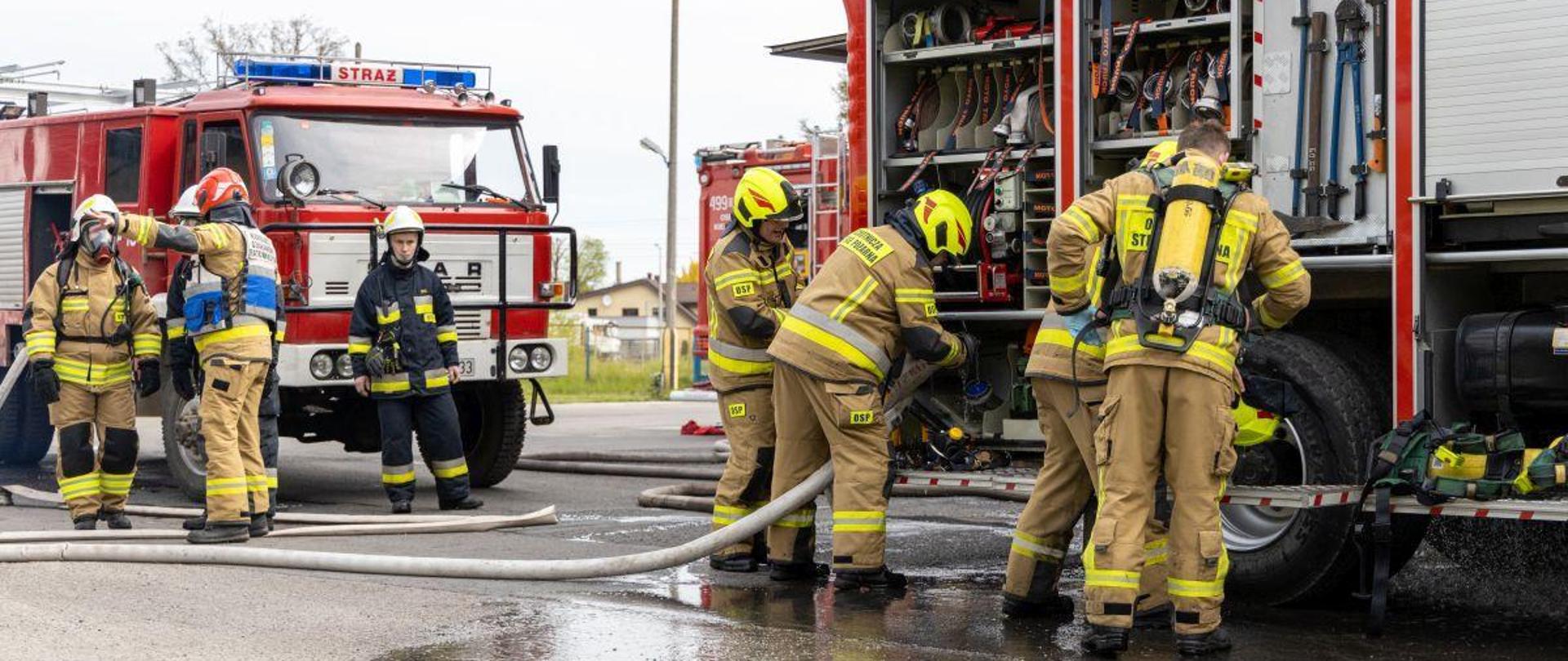 Na zdjęciu znajdują się strażacy w ubraniach specjalnych oraz samochody strażackie do których strażacy podłączają węże