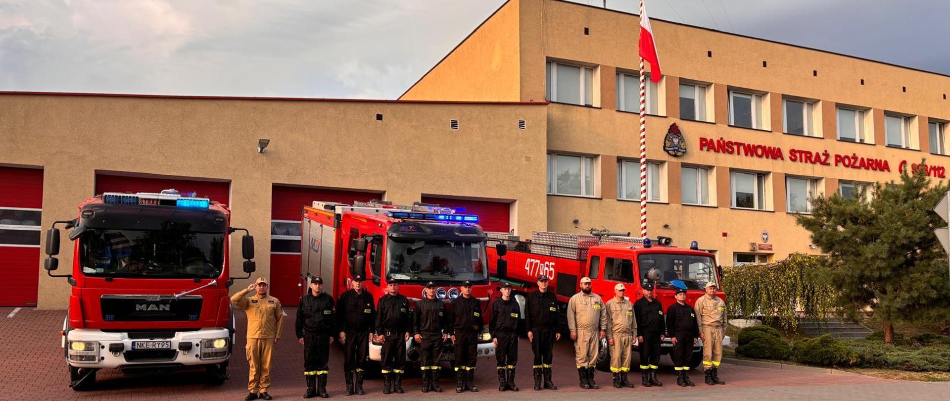 Strażacy w mundurach służbowych i koszarowych oddają hołd tragicznie poległym strażakom. W tle czerwone samochody strażackie oraz budynek komendy.