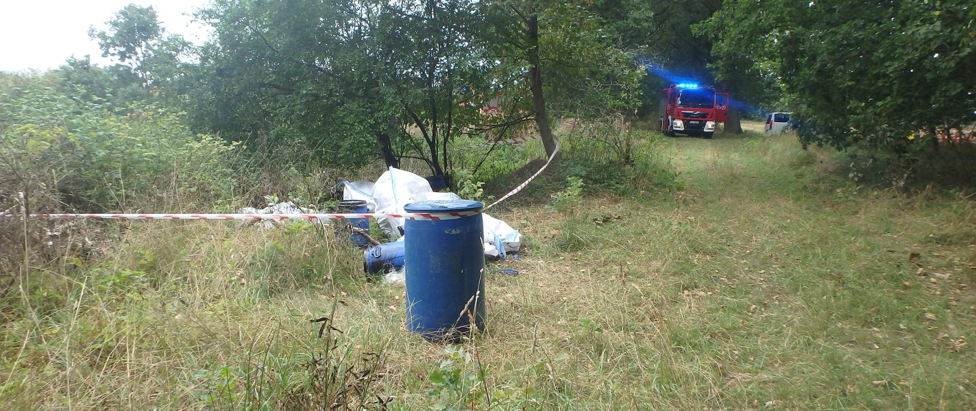 Zdjęcie przedstawia porzucone odpady w postaci beczek wypełnionych czarna mazistą substancją. Beczki znajdują się bezpośrednio na gruncie, naturalnym podłożu. Na zdjęciu widać drogę leśną, nieutwardzoną ścieżkę oraz teren porośnięty przez wielogatunkową roślinność. W tle widać wóz Straży Pożarnej oraz samochód Wojewódzkiego Inspektoratu Ochrony Środowiska.