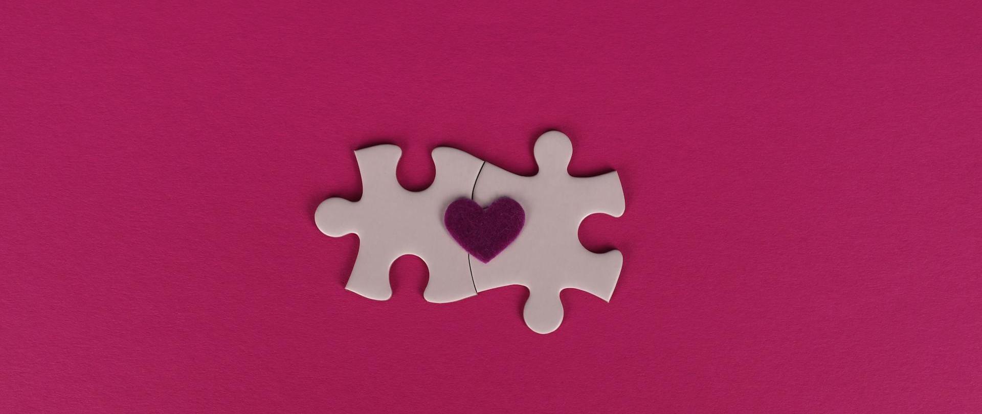Zdjęcie przedstawia dwa połączone różowe puzzle leżące na rubinowym tle. Na ich wierzchu położone jest małe bordowe serduszko.