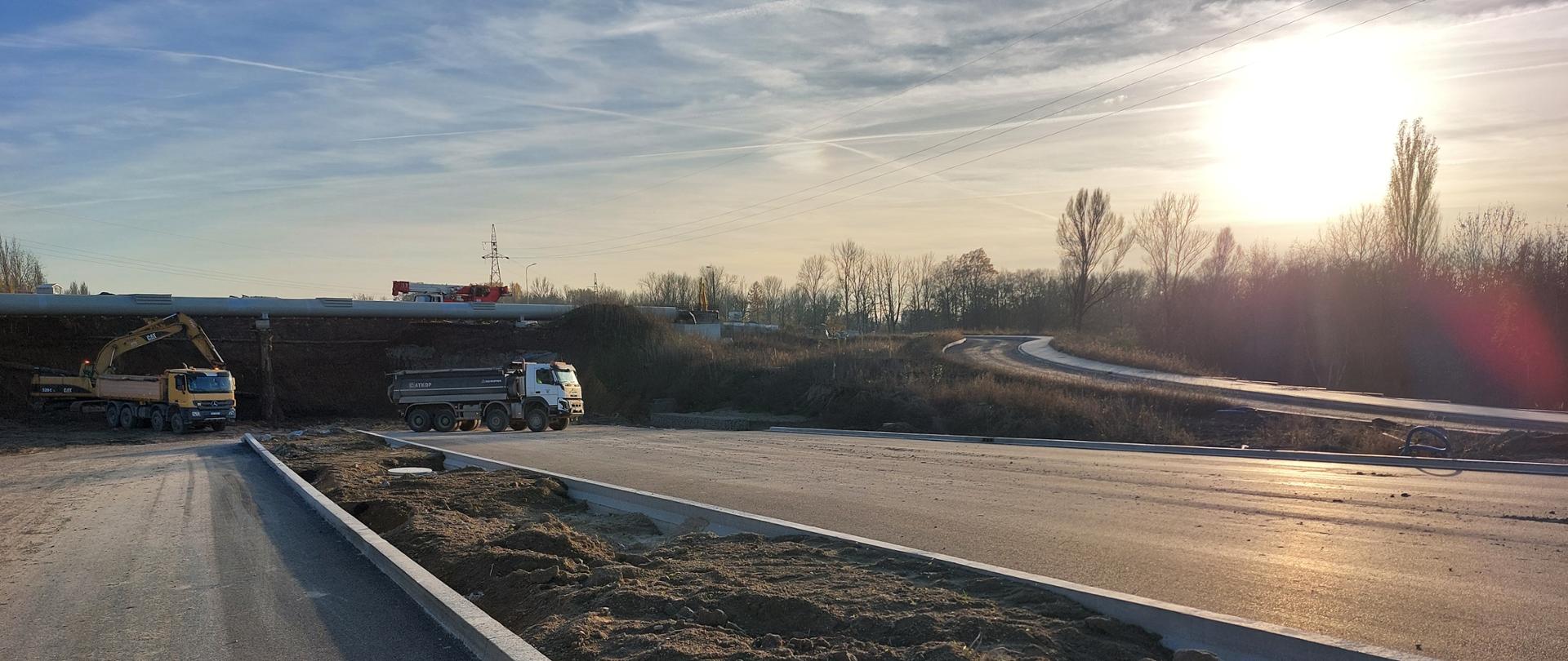 Zdjęcie zrobione z poziomu ziemi. Na pierwszym planie widoczne jezdnie z pasem rozdzielającym oraz ciężki sprzęt. 