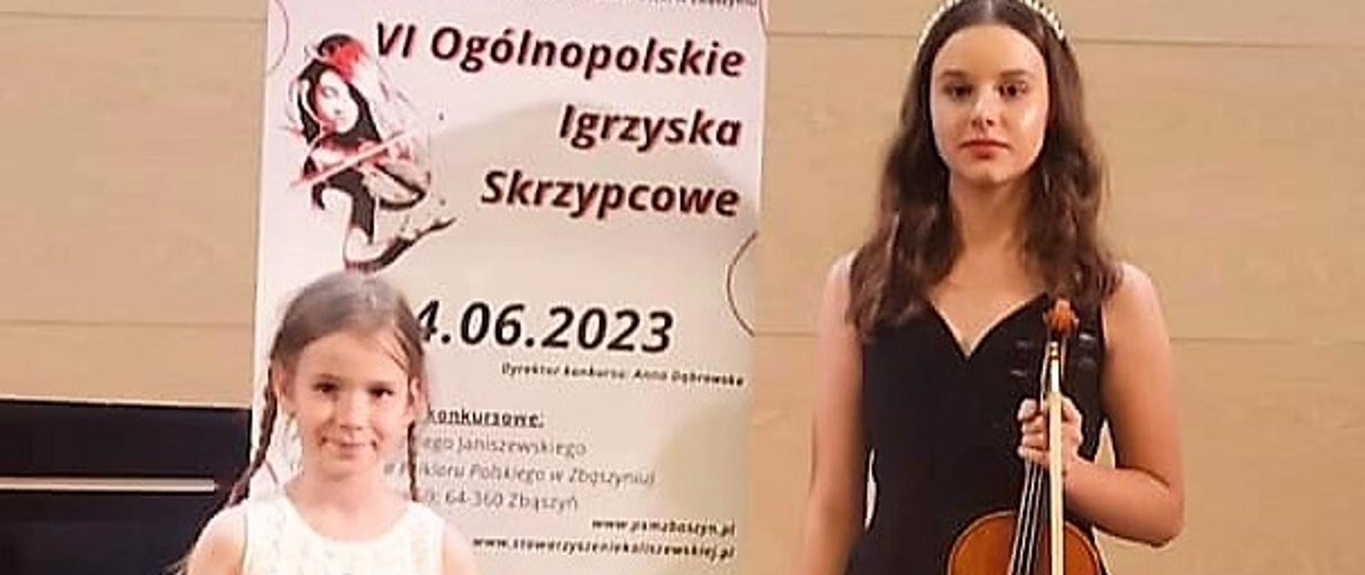 dwie dziewczynki ze skrzypcami, pomiędzy nimi plakat na sztaludze, za nimi beżowe tło