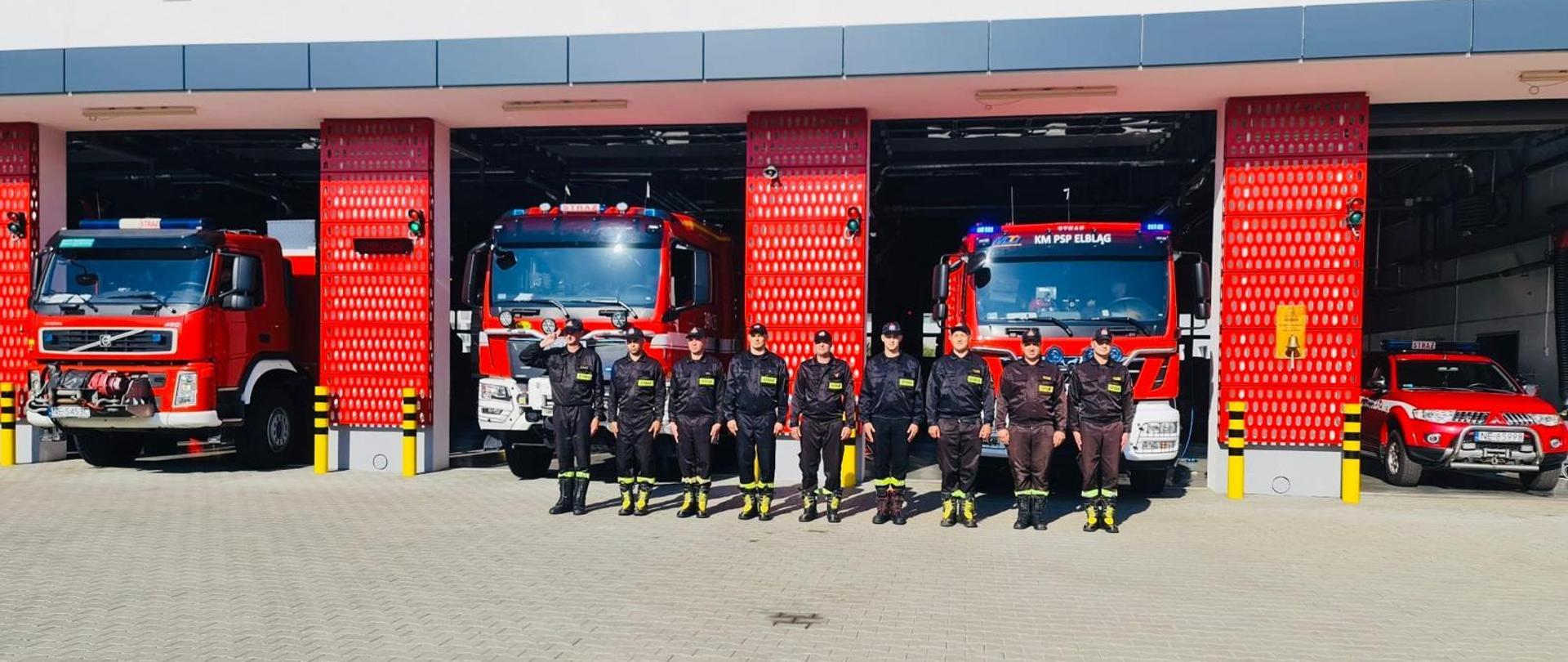 Na zdjęciu znajduje się 9 strażaków, którzy stoją w szeregu. Strażacy oddają hołd powstańcom poległym w 80 rocznicę wybuchu Powstania Warszawskiego. Strażacy mają na sobie mundury koszarowe w kolorze czarnym. W tle stoją pojazdy pożarnicze