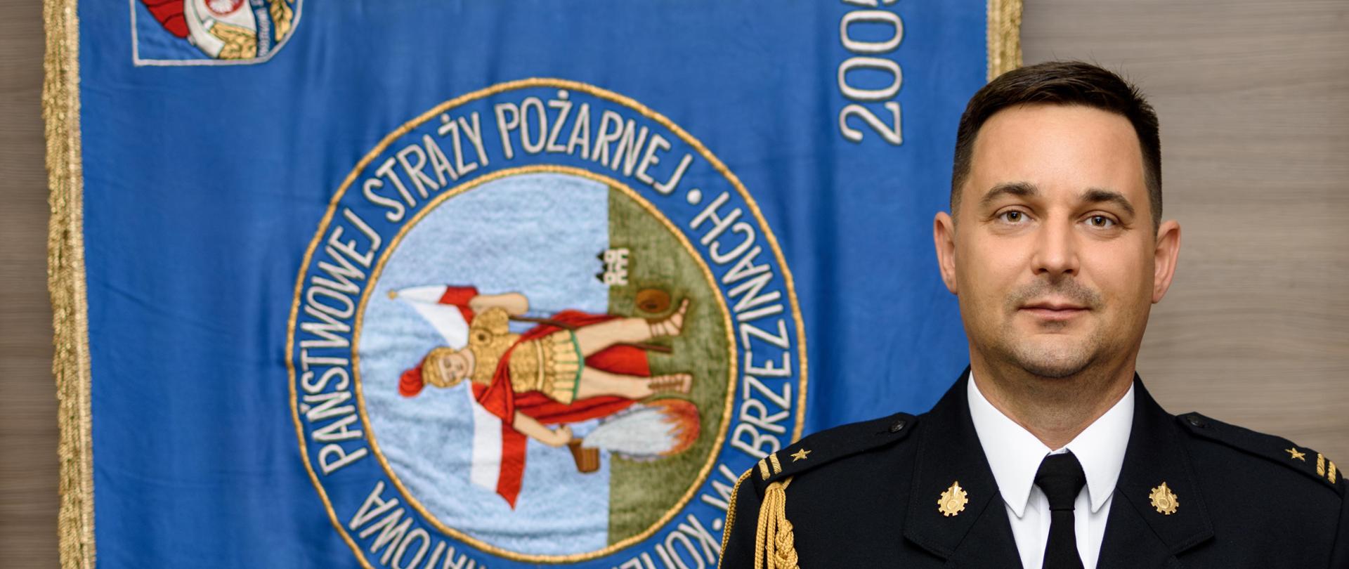 Zdjęcie profilowe zastępcy Komendanta Powiatowego na tle sztandaru Komendy Powiatowej PSP w Brzezinach