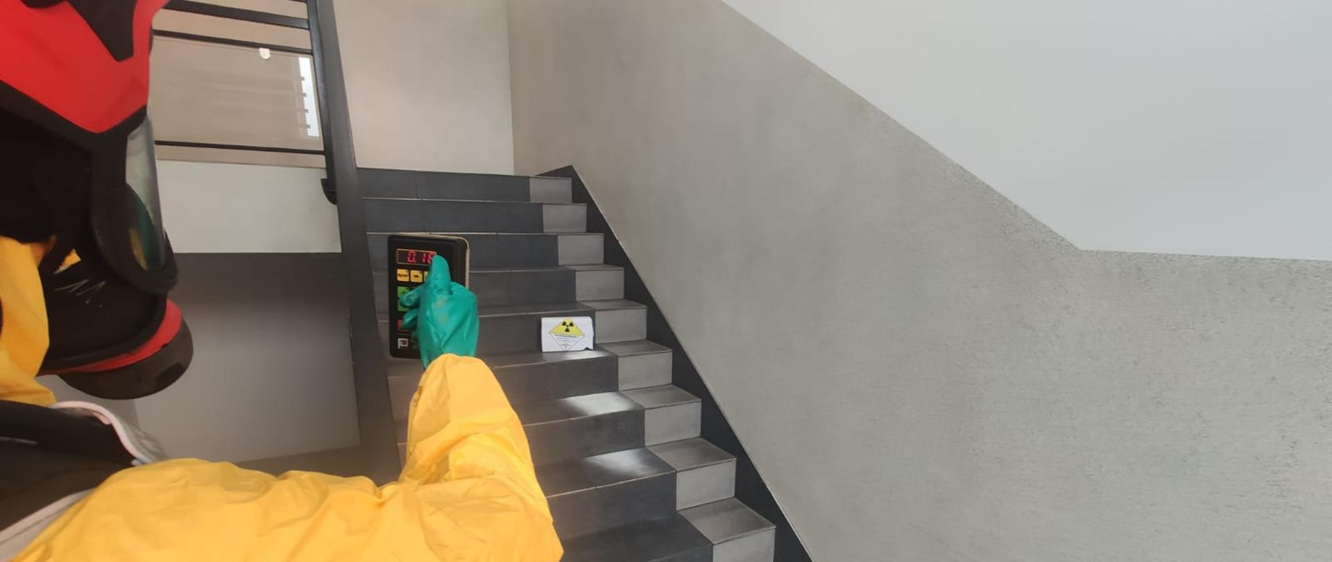 Strażak w ubraniu przeciwchemicznym dokonuje pomiaru promieniowania. Na schodach stoi uszkodzona paczka z czarną koniczyną na żółtym trójkącie. Poniżej rozsypany biały praoszek.