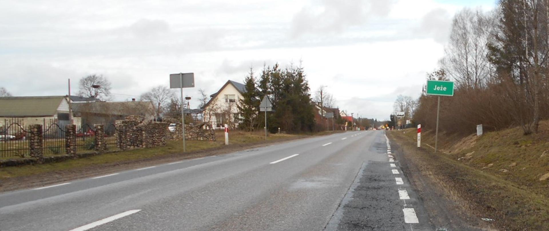 DK63 Borki - Jeże. Wjazd do miejscowości Jeże. Po lewej stronie drogi zabudowania, po prawe znak z nazwą miejscowości.