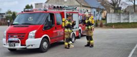 Teren firmy branży mięsnej. Na placu manewrowym stoją dwa lekkiei samochoy pożarnicze. Obok nich znajduje się dwóch strażaków, którzy ubierają maski twarzowe aparatów powietrznych. Strażacy mają na sobie ubrania specjalne w kolorze piaskowym.W tle budynki i drzewa. 