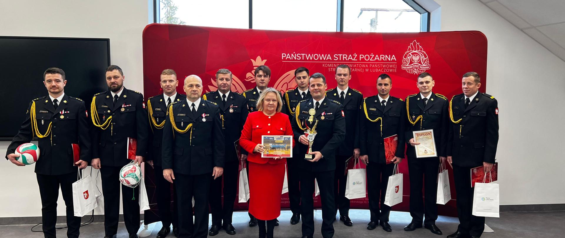 Zdjęcie przedstawia funkcjonariuszy PSP w mundurze wyjściowym oraz kobietę w czerwonej sukience (Pani Starosta Barbara Broź). W tle widoczna czerwona ścianka z napisem Komenda Powiatowa PSP w Lubaczowie. 