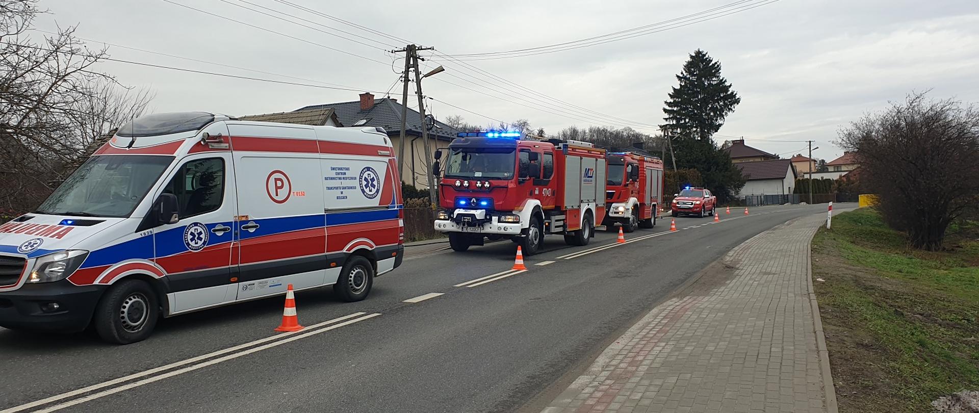 Strażackie samochody stoją na jednym pasie drogi z włączonymi sygnałami świetlnymi, Przed nimi karetka pogotowia ratunkowego. Kliknięcie powoduje powiększenie.