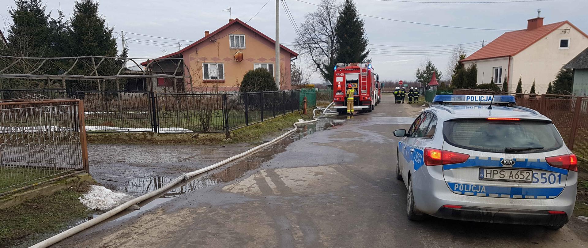 Na zdjęciu widoczne są stojące na jezdni pojazdy. Z prawej radiowóz Policji, w oddali z lewej samochód Państwowej Straży Pożarnej z rozwiniętą linią zasilającą. Obydwa pojazdy mają włączone sygnały świetlne. W oddali widać ratowników podczas działań ratowniczo-gaśniczych.