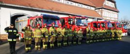 Strażacy Komendy Miejskiej Państwowej Straży Pożarnej w Zabrzu stoją w szeregu przed wystawionymi z garażu Komendy samochodami strażackimi z włączonymi sygnałami błyskowymi.