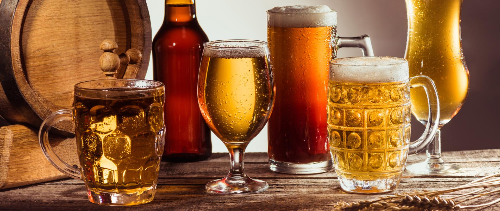 Na zdjęciu jest drewniana beczka wraz z piwem znajdującym się w różnych szklanych kubkach i szklankach oraz w butelce.