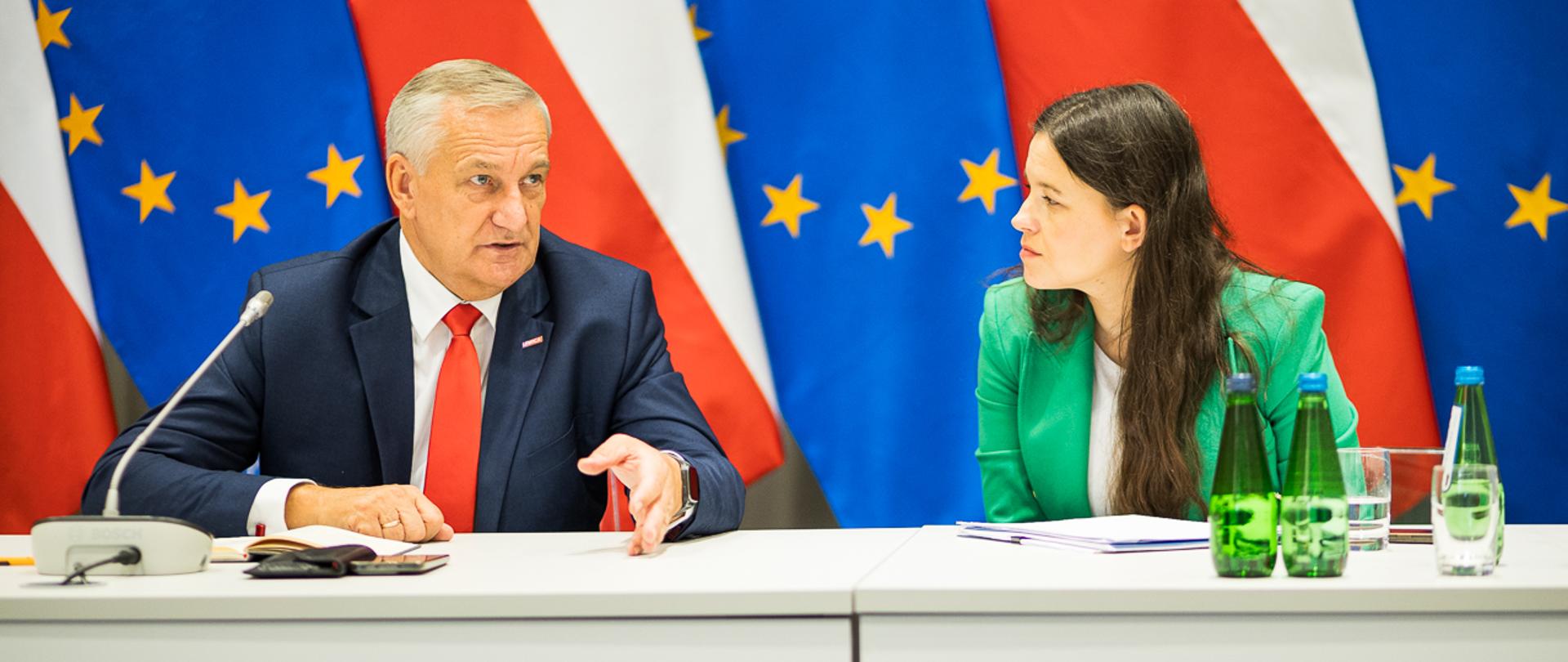 Mężczyzna i kobieta za stołem prezydialnym na tle flag Polski i UE.