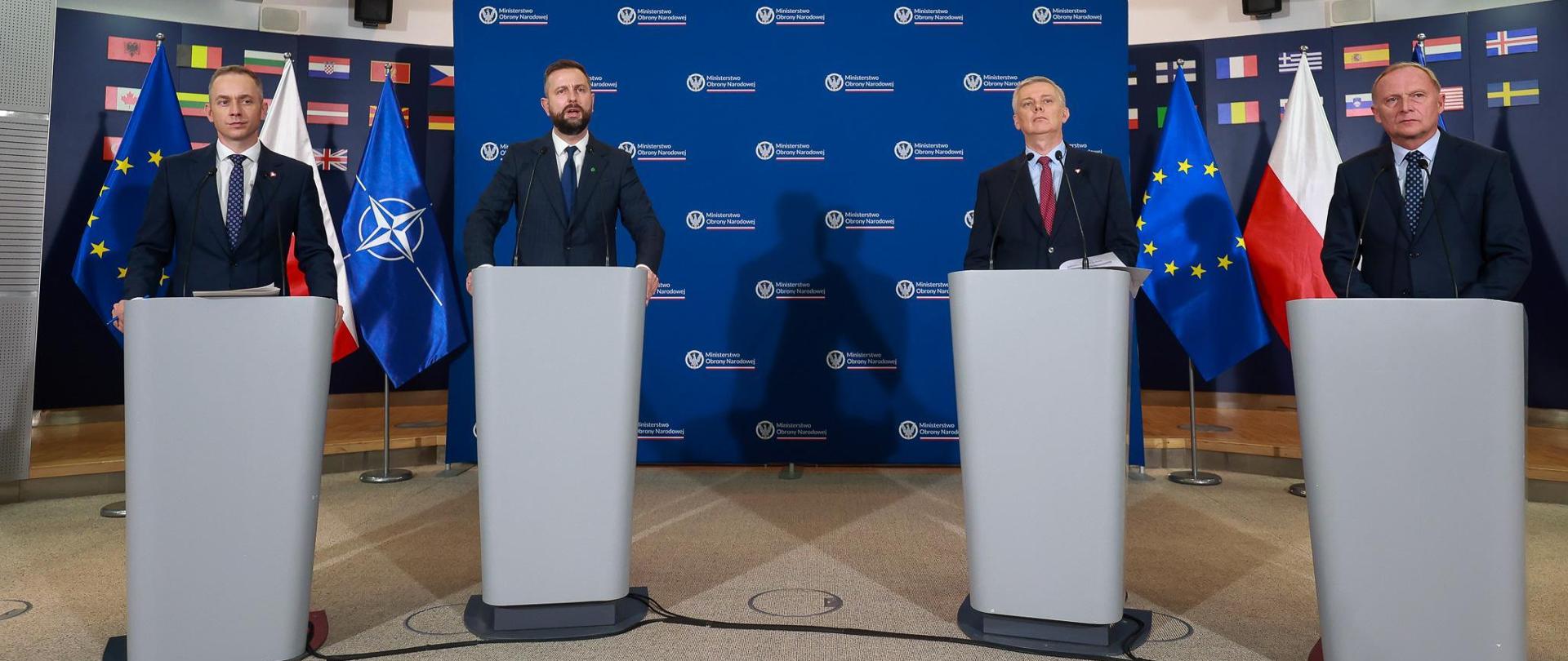 Czterech mężczyzn w ciemnych garniturach przed mównicami z mikrofonami. W głębi na tle granatowej ściany flagi Polski, Unii Europejskiej i NATO.