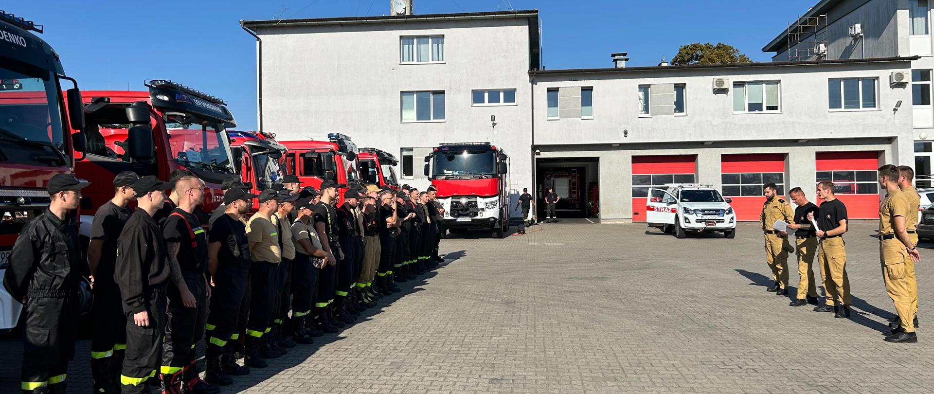 Fotografia przedstawia ujęcie z boku pododdziału kompanii gaśniczej. Przed pojazdami pożarniczymi zaparkowanymi w rzędzie stoją strażacy w dwuszeregu, gotowi do podjęcia działań przeciwpowodziowych. Naprzeciwko kompanii widoczna kadra dowódcza Państwowej Straży Pożarnej prowadząca odprawę związku taktycznego. W tle widoczny budynek Komendy Powiatowej Państwowej Straży Pożarnej w Strzelcach Krajeńskich.