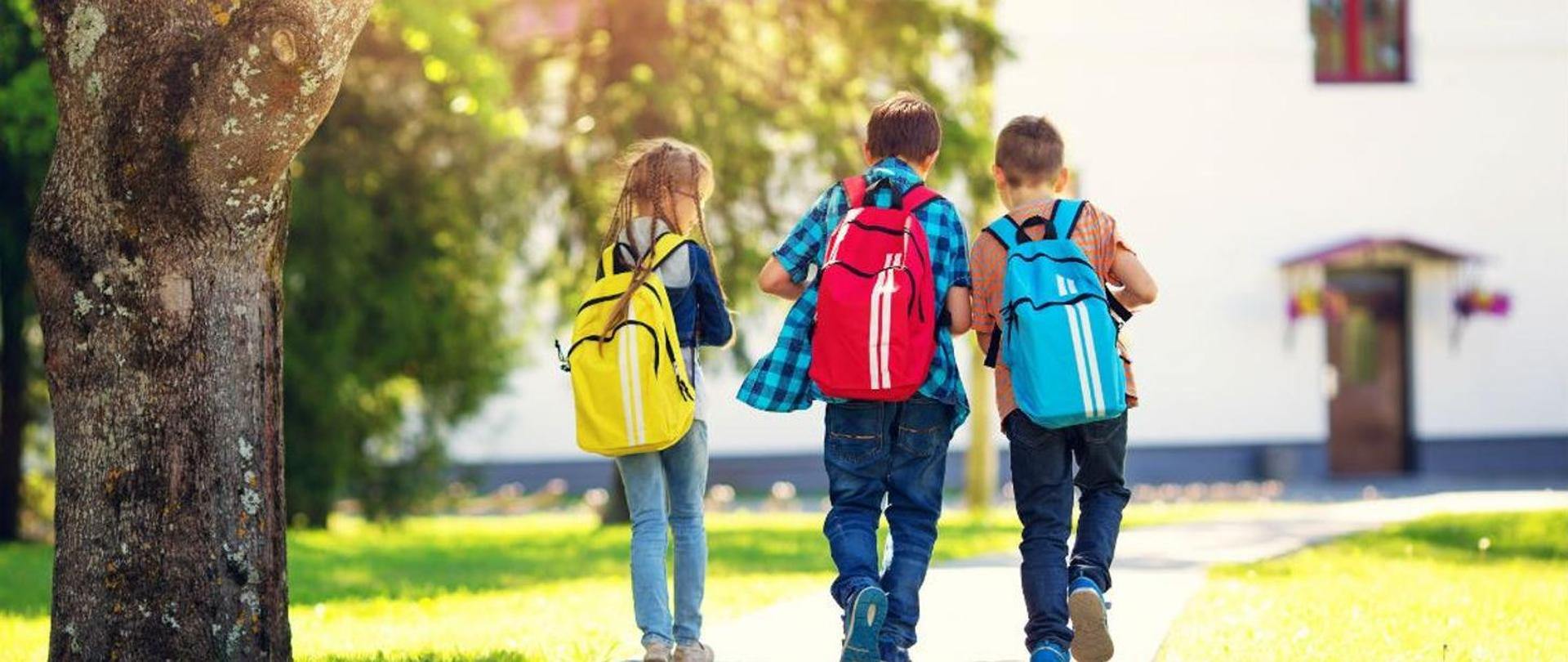 Zdjęcie przedstawiające trójkę dzieci niosących na plecach plecaki szkolne.