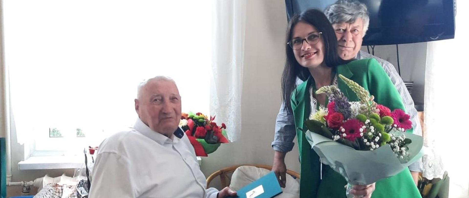 Uśmiechnięta kobieta trzyma w ręce bukiet kwiatów, za nią stoi mężczyzna, przed nimi siedzi starszy mężczyzna z ozdobną teczkę z logo KRUS w rękach