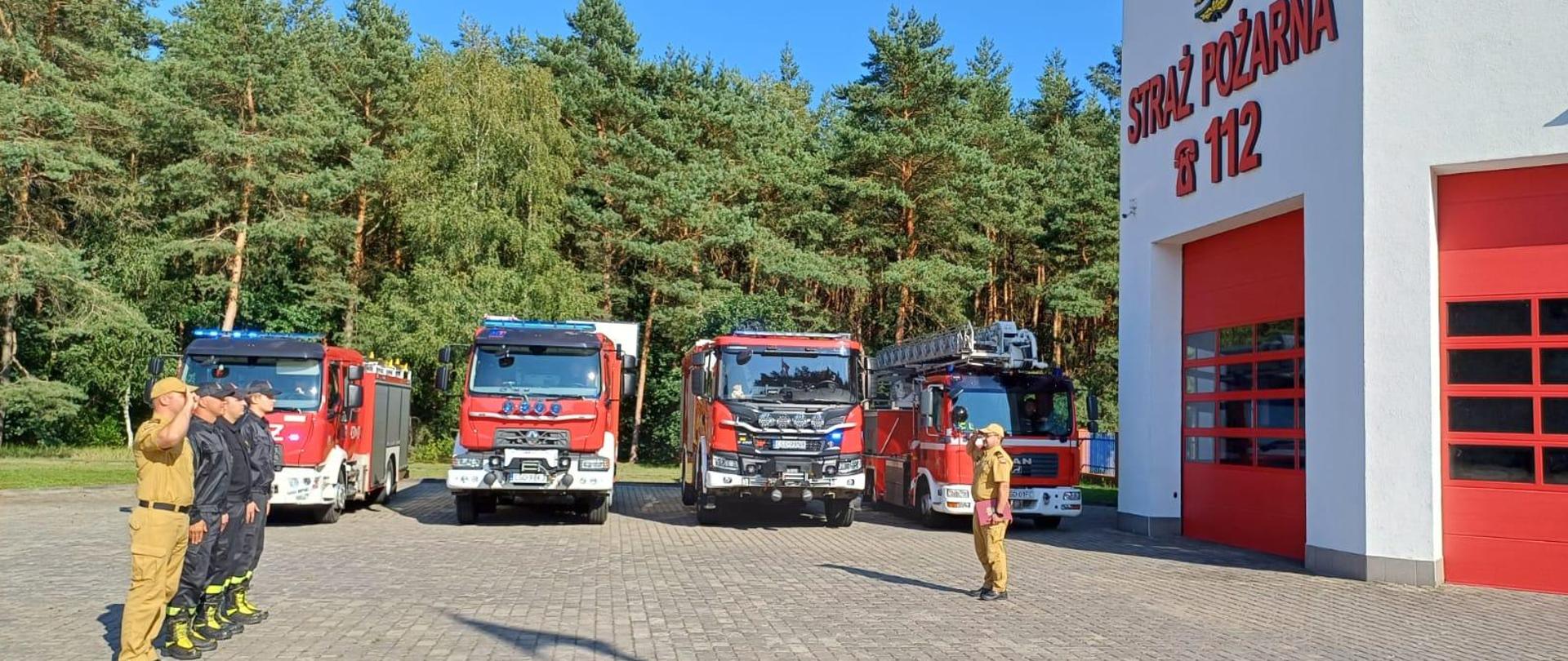 Komendant Powiatowy oraz Zastępca Dowódcy JRG salutują a pozostali funkcjonariusze stoją w szeregu na baczność. W tle widać pojazdy pożarnicze.