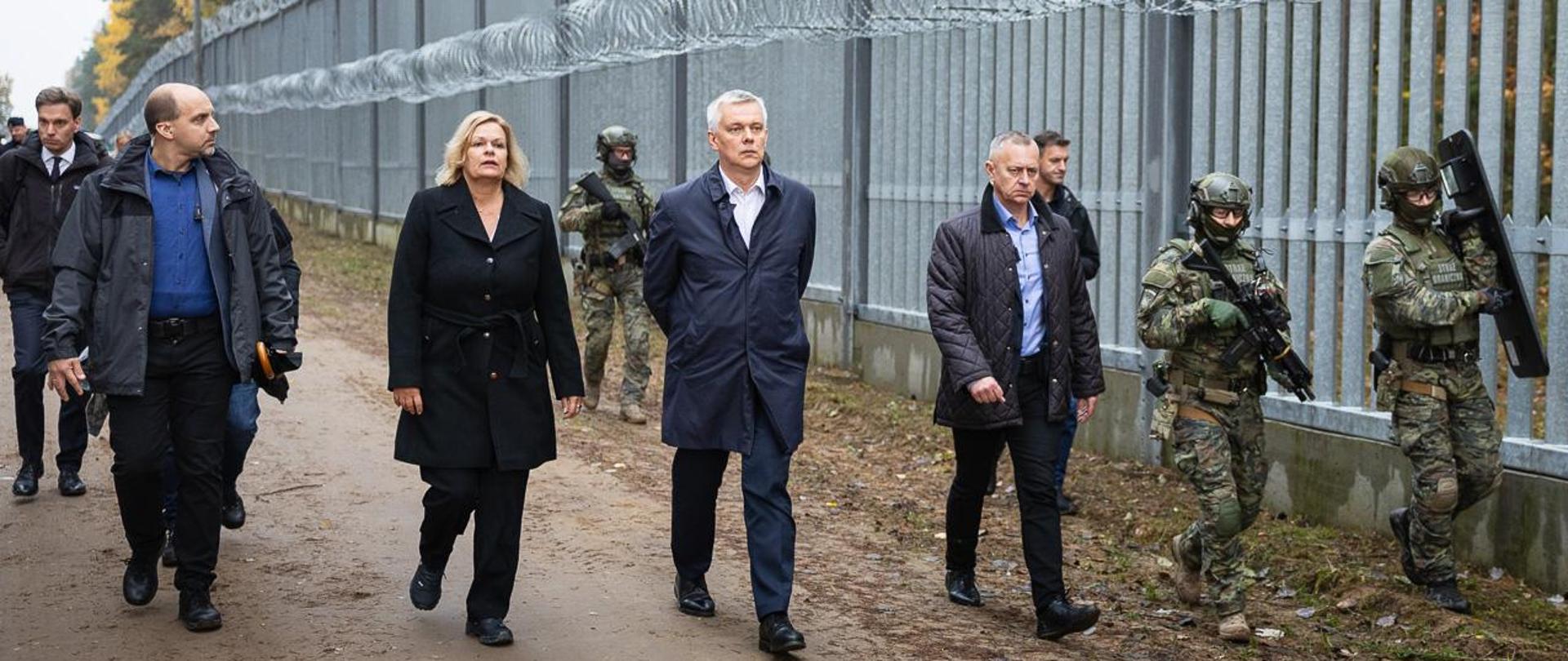 Ministers Tomasz Siemoniak and Nancy Faeser visit the strip on the Polish-Belarusian border. They are accompanied by a group of people. The barrier is guarded by armed border guards
