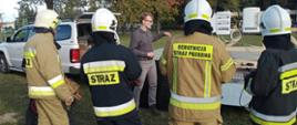 Teren stadionu w Rawiczu. Na przyczepce samochodowej ustawiony jest model instalacji fotowoltaicznej. Obok przyczepki stoi instruktor, który przedstawia budowę i zasadę działania instalacji. O przyczepkę oparte są panele fotowoltaiczne. Na przeciwko przyczepki znajduje sie 5 uczestników warsztatów w ubraniah specjalnych, ktorzy słuchają wykładu. 