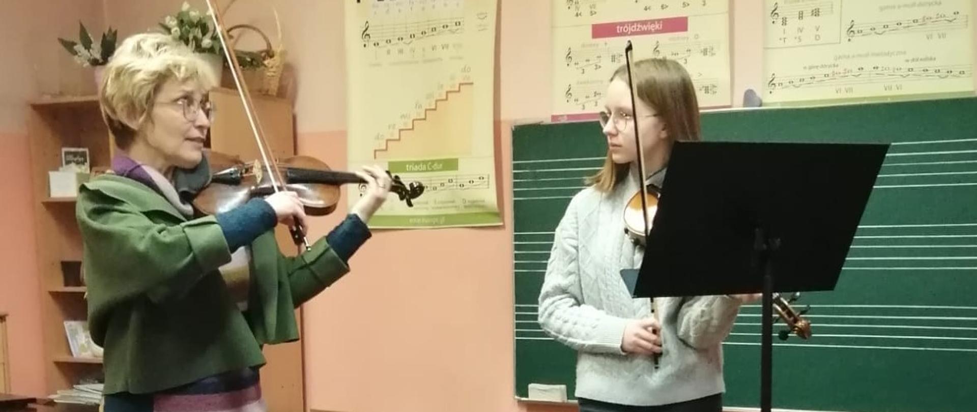 Prowadząca pokazująca na skrzypcach technikę. obok uczennica obserwująca prezentację