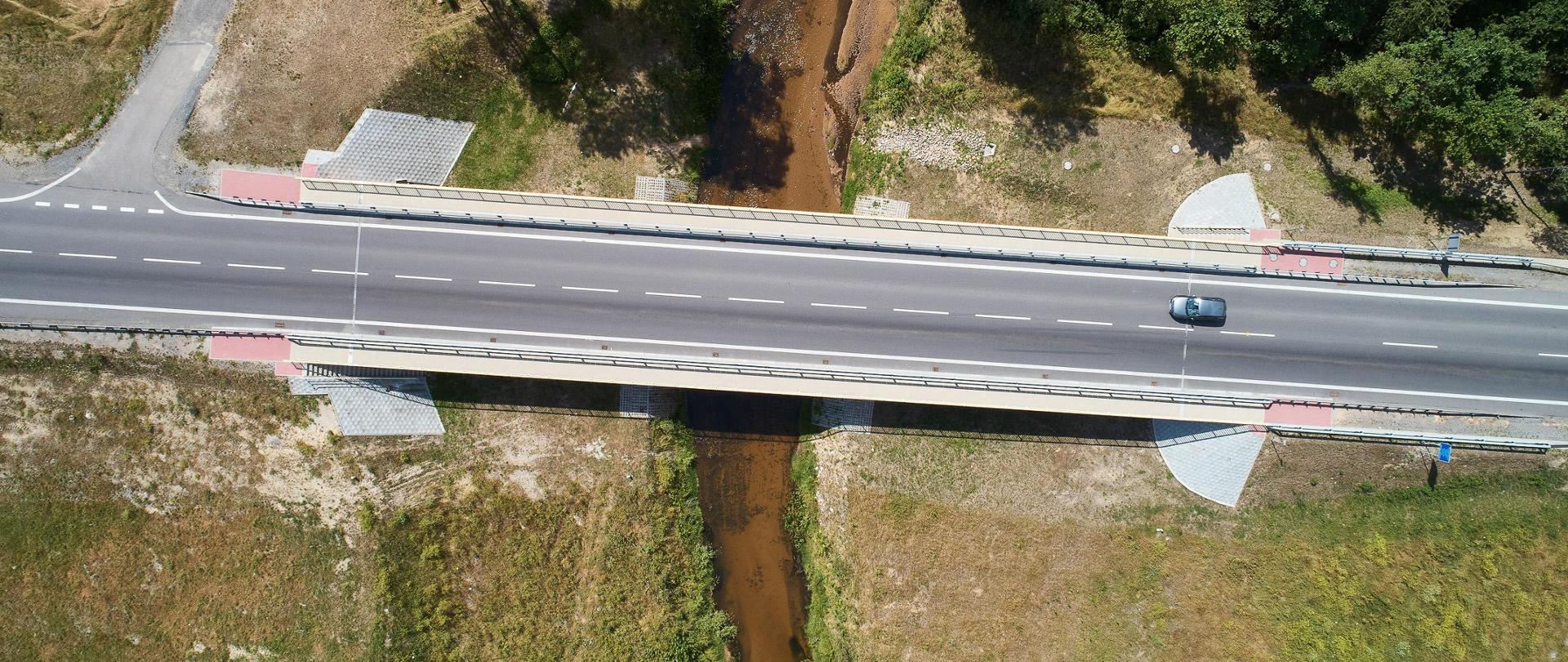 Widok z góry na drogę krajową nr 19 w Domostawie. Na środku widać drogę z mostem, która przebiega nad rzeką. Dookoła drogi zieleń, trawa, drzewa, krzewy. Na drodze widać auto osobowe. 