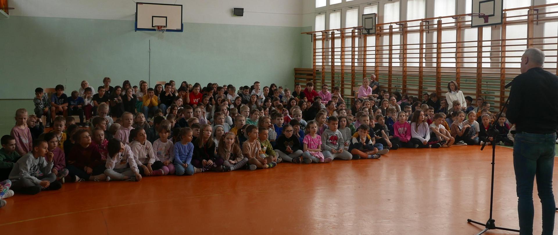 Sala gimnastyczna i dzieci siedzące na podłodze, słuchające zapowiedzi koncertu przez konferansjera.