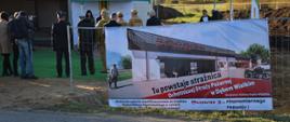 Widok na teren budowy OSP w Dębem Wielkim, na pierwszym planie duży plakat zawieszony na ogrodzeniu z napisem TU POWSTAJE STRAŻNICA OCHOTNICZEJ STRAŻY POŻARNEJ W DĘBEM WIELKIM, na drugim planie zebrani goście pod budynkiem strażnicy