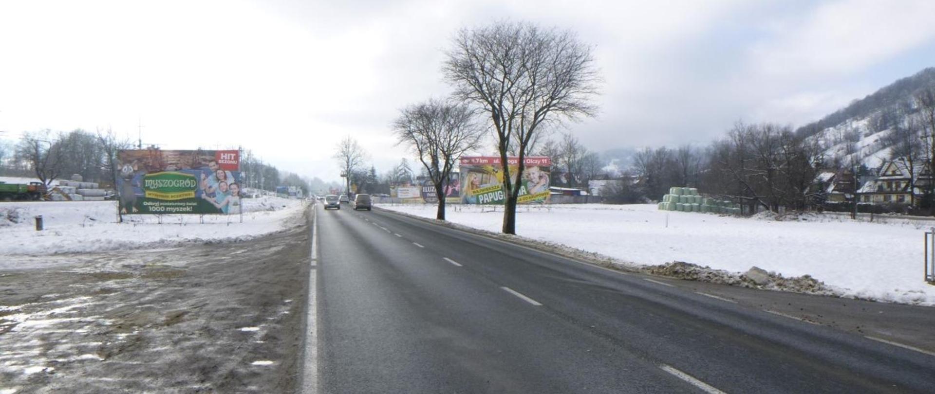 Zima. Zdjęcie z poziomu ziemi. Widoczne pojazdy jadące jednojezdniową drogą. Na poboczach śnieg i bilbordy reklamowe.