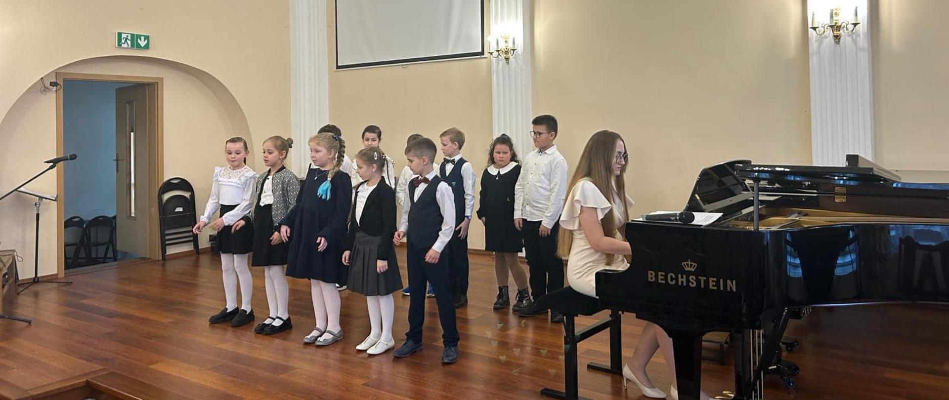Uczniowie klasy II/6 na scenie auli podczas popisu wraz z nauczycielką grająca na fortepianie.