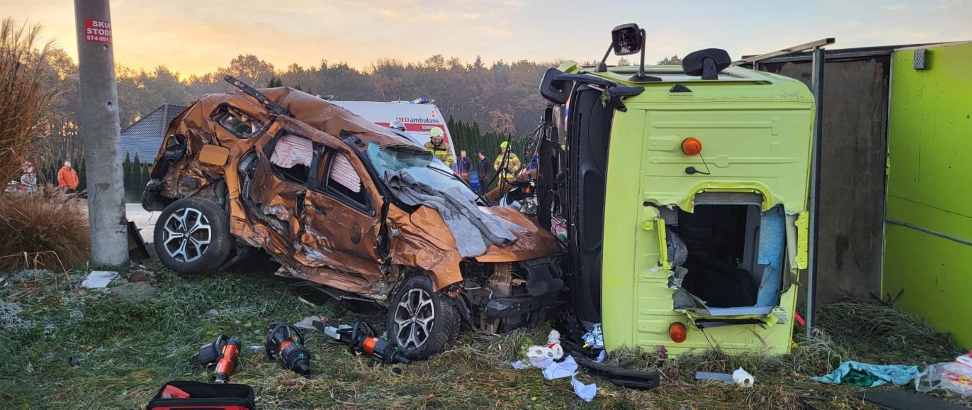 Zdjęcie przedstawia wypadek samochodu osobowego z ciężarowym. Na zdjęciu po lewej stronie widać uszkodzony całą prawą stronę, maskę i dach samochodu osobowego w kolarze brązowym, samochód ma rozbite szyby. Po prawej stronie przy samochodzie osobowym leży przewrócony na lewy bok samochód ciężarowy, który ma rozcięty przez strażaków dach w celu dostania się do poszkodowanego kierowcy. Przed pojazdami leżą na ziemi hydrauliczny sprzęt do cięcia i rozpierania , torba medyczna oraz rzeczy które wypadły z kabiny pojazdów. Za pojazdami widać część pojazdu Zespołu Ratownictwa Medycznego a obok nich stoją strażak ubrany w piaskowe ubranie specjalne na głowie ma hełm, za nim stoją osoby postronne.