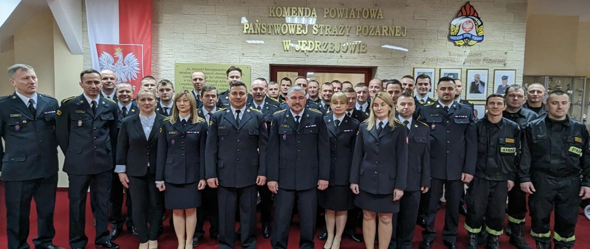 Funkcjonariusze i pracownicy Komendy Powiatowej Państwowej Straży Pożarnej w Jędrzejowie na uroczystej zbiórce pożegnania emerytów. Wszyscy znajdują się na sali tradycji.