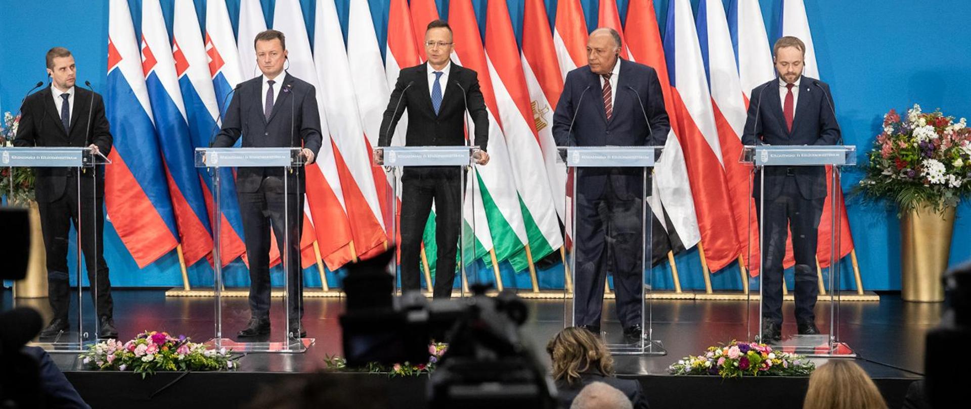 Minister Mariusz Błaszczak wraz z innymi ministrami stoją przy mównicach.