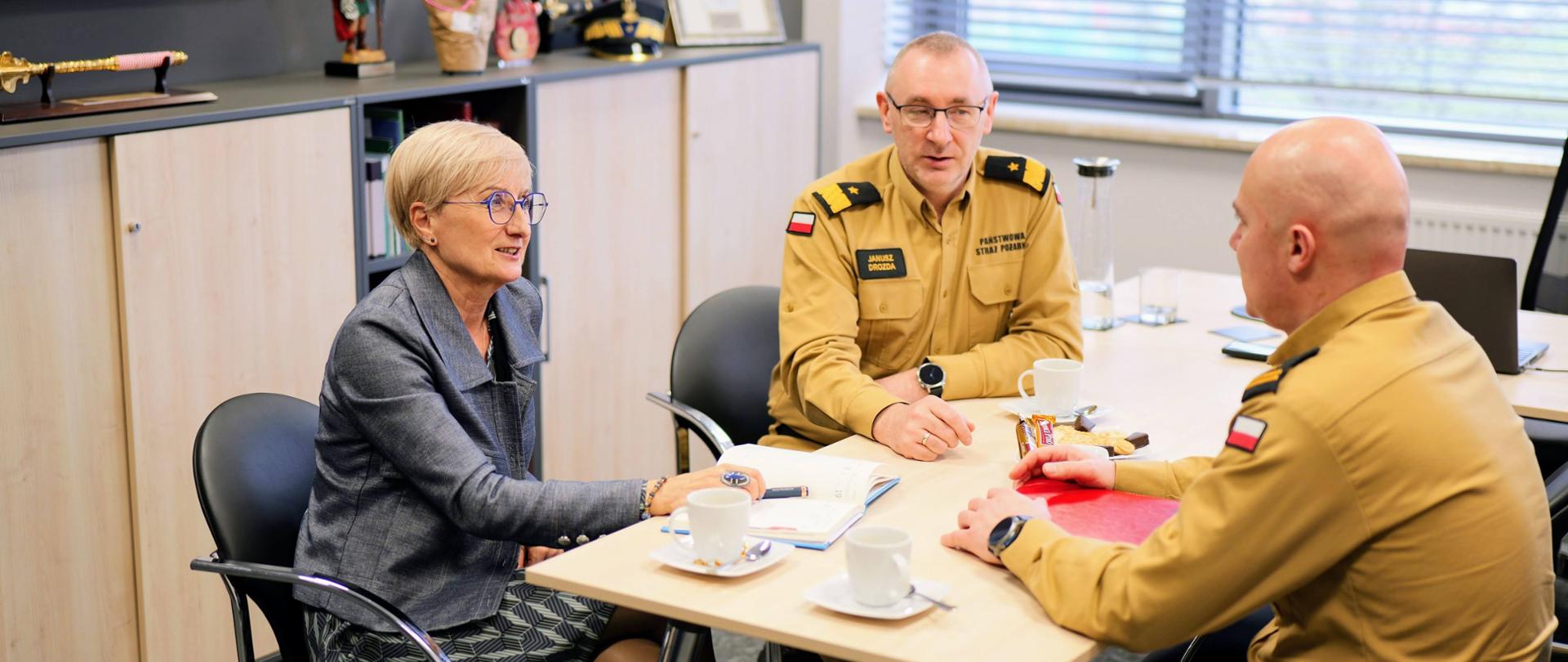 Przy prostokątnym stole w pomieszczeniu biurowym siedzą trzy osoby. Kobieta z krótkimi, jasnymi włosami i w okularach (starosta żagański) ma przed sobą notatnik. Dwaj mężczyźni (po lewej komendant wojewódzki, na prawo komendant powiatowy w Zaganiu), ubrani w jasnobrązowe mundury strażackie, siedzą obok i słuchają jej wypowiedzi. Na stole stoją białe filiżanki z kawą i talerzyki z przekąskami. W tle widać jasne szafki oraz figurki dekoracyjne.