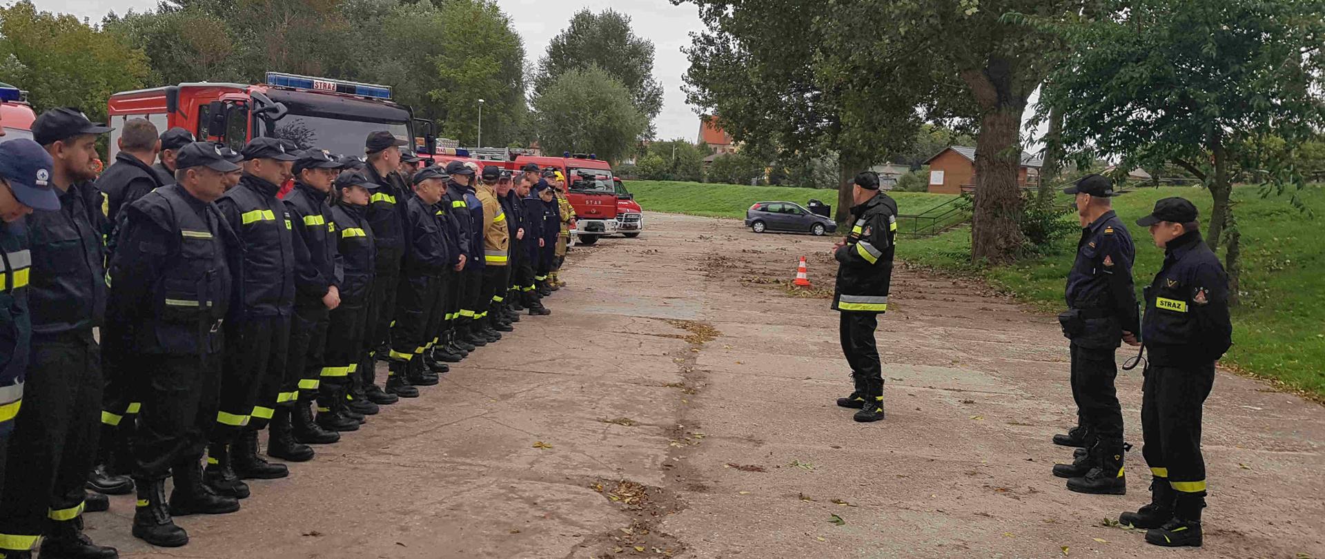 Na zdjęciu widać stojących strażaków na zbiórce i za nimi samochody. Zdjęcie zrobione w dzień. 